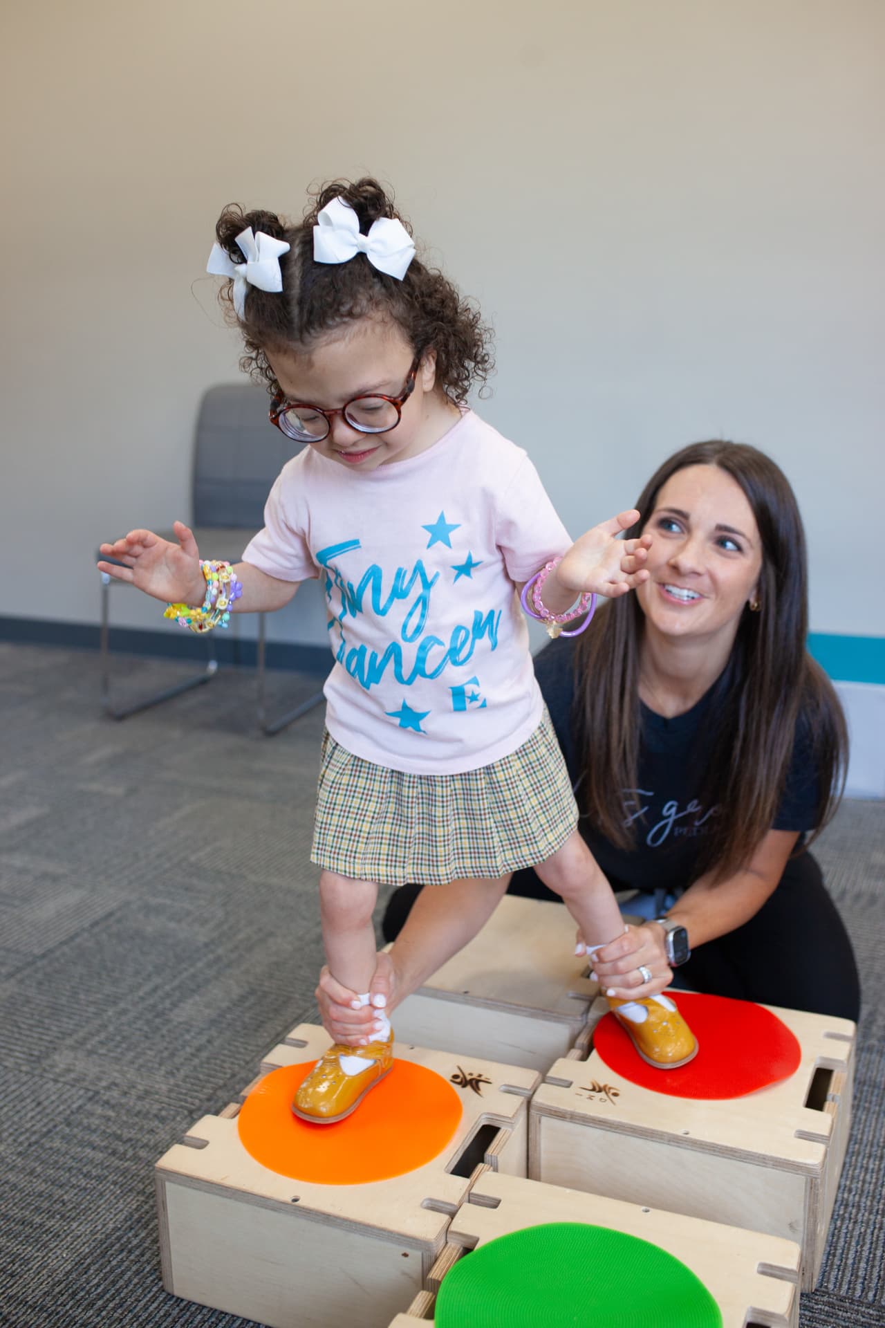 A pediatric therapist supports a young child with white bows as she stands on colorful DMI stepping platforms.