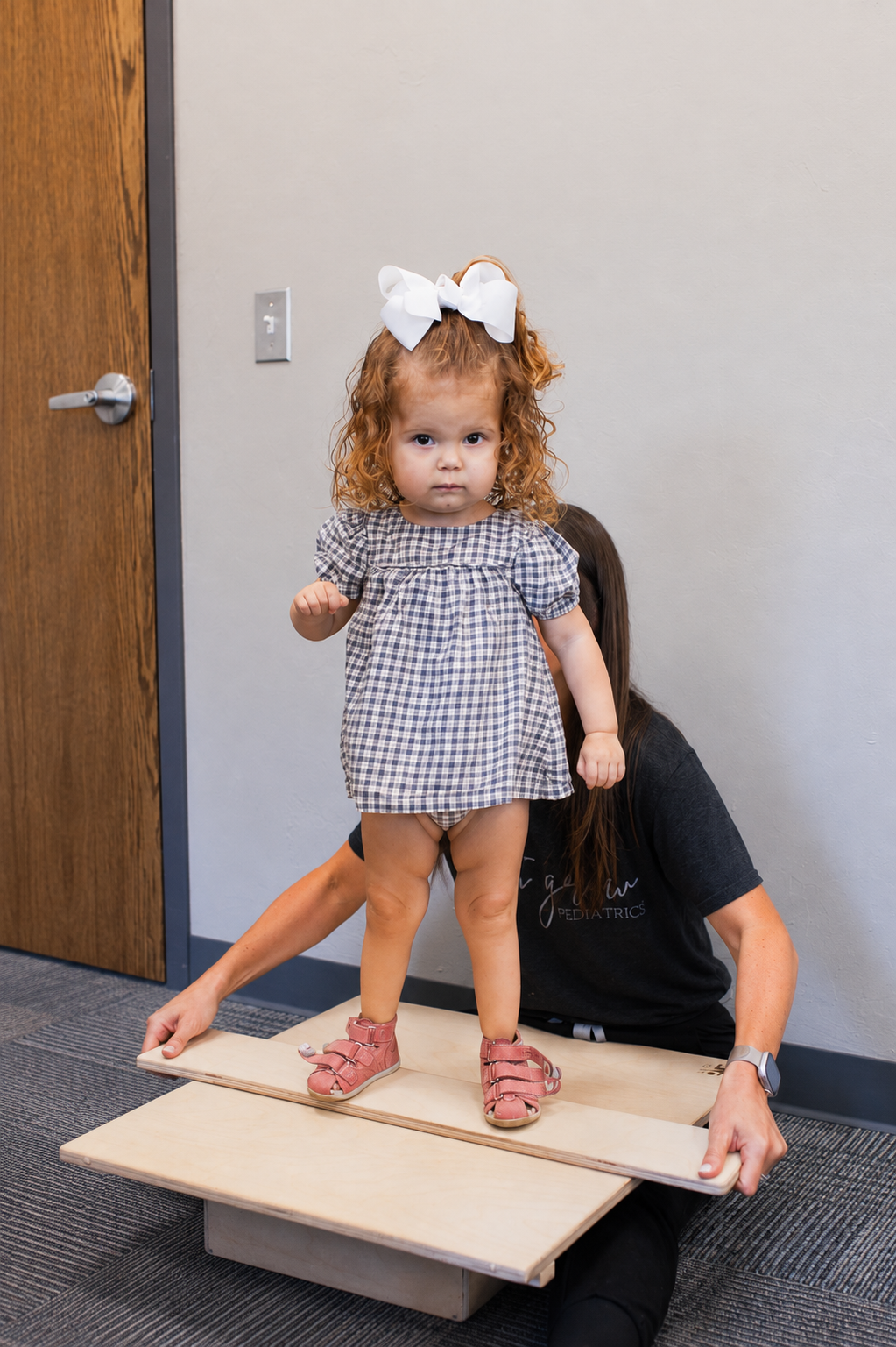 A pediatric therapist supports a young child as they stand on a wooden balance board, focusing on balance and confidence through guided movement.