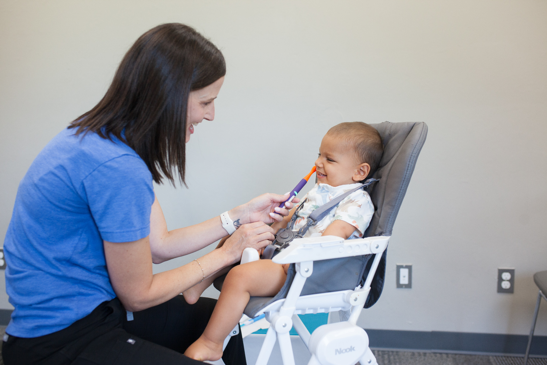 A pediatric occupational therapist smiles while feeding a baby with a colorful spoon during a calm feeding therapy session.