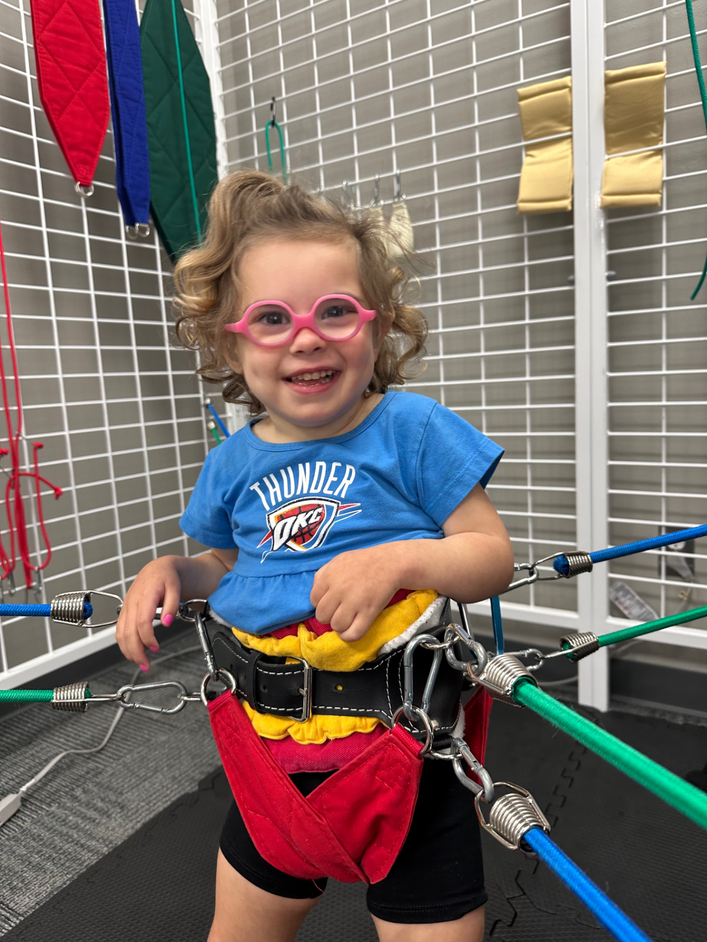 A smiling child wearing pink glasses supported by a therapy harness.