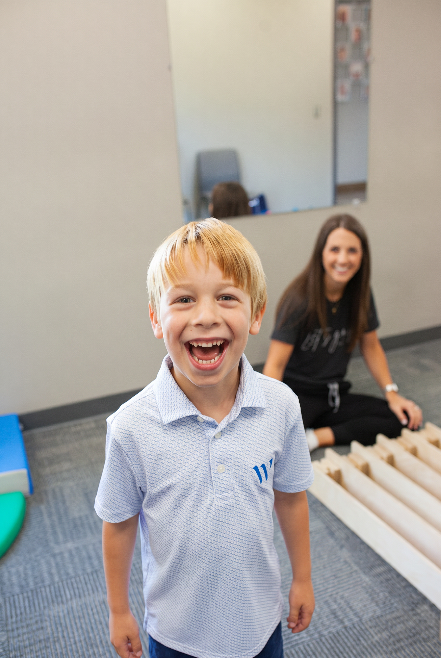 A smiling child standing confidently in the therapy gym with a pediatric therapist seated nearby