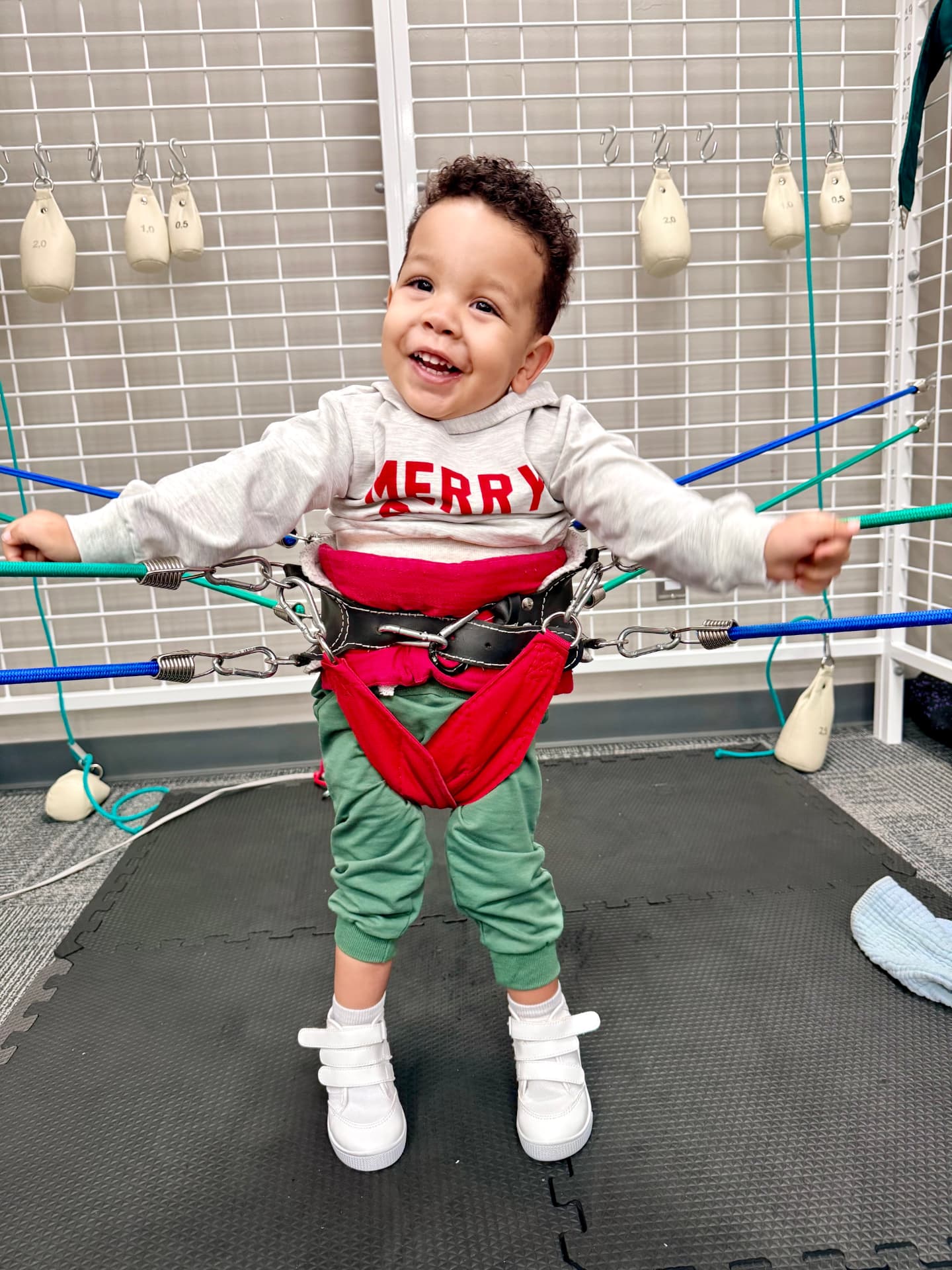 A smiling toddler standing supported by bungee cords in the universal exercise unit.