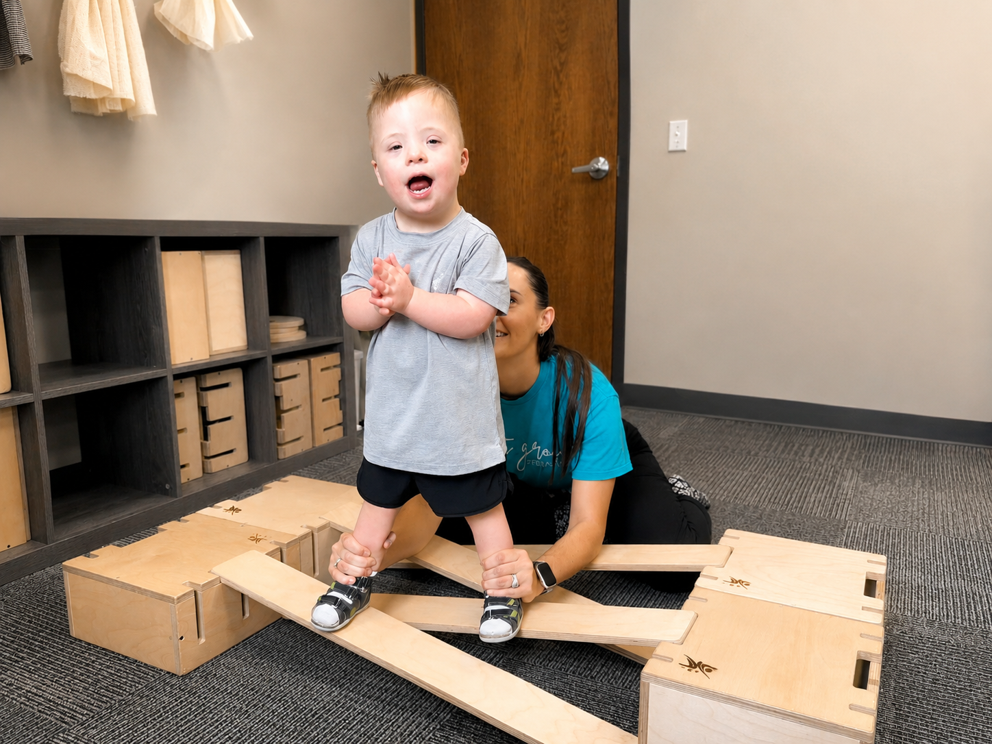 A young child standing with therapist support on wooden balance equipment during an intensive therapy session.